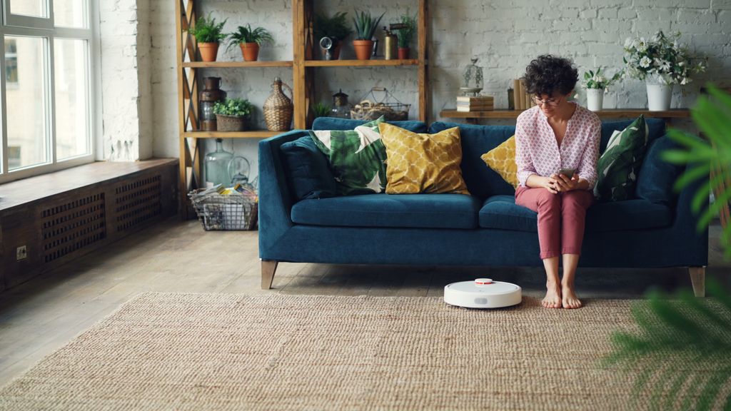 A woman sits on a sofa in a cozy living room with a robot vacuum on the floor.